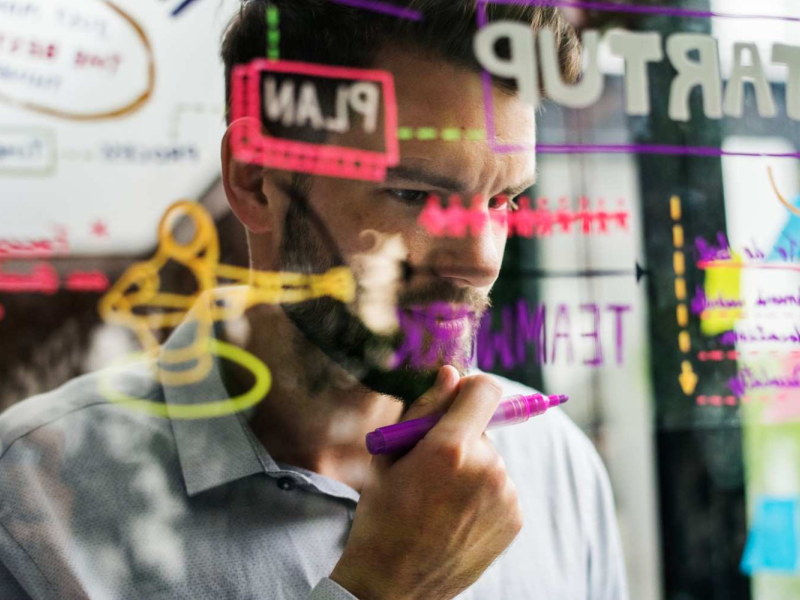 Profissional escrevendo em vidro transparente com anotações coloridas de planejamento.