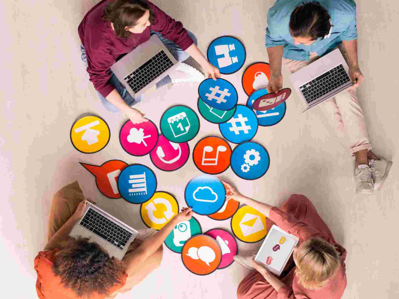 Equipe colaborando em torno de ícones digitais representando funcionalidades de produtos, com notebooks e ambiente criativo.